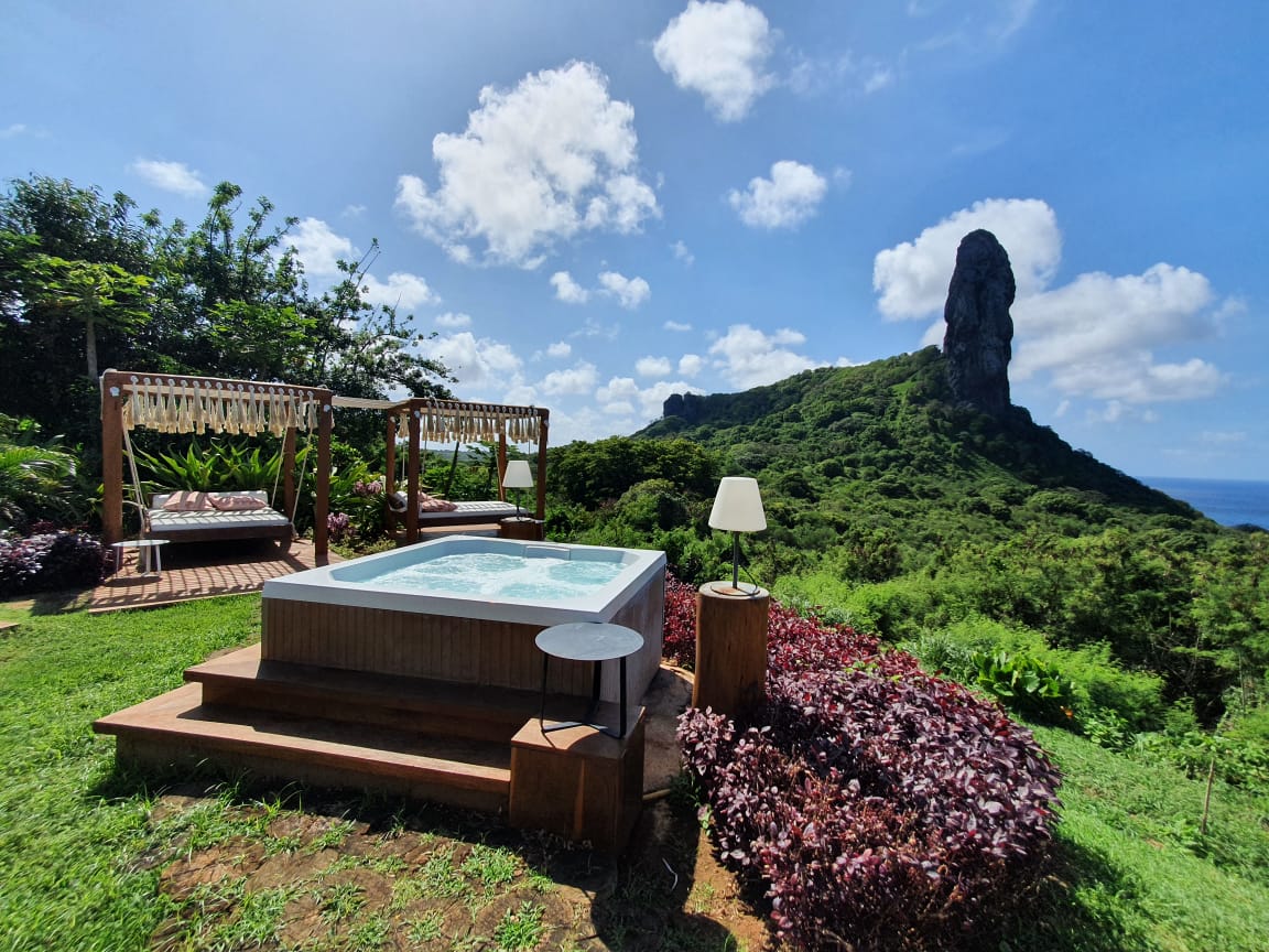 Planejar uma viagem à Fernando de Noronha. A imagem mostra uma piscina quadrada no alto de uma montanha, com tendas ao lado. Ao fundo, paisagem de montanhas e céu azul.