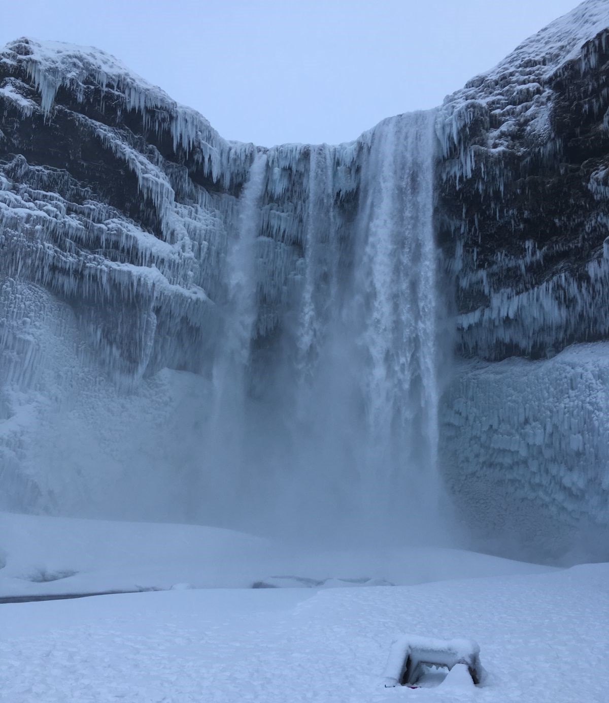 Cenário mostra como é a Islândia: uma cachoeira alta em meio ao solo coberto de neve.